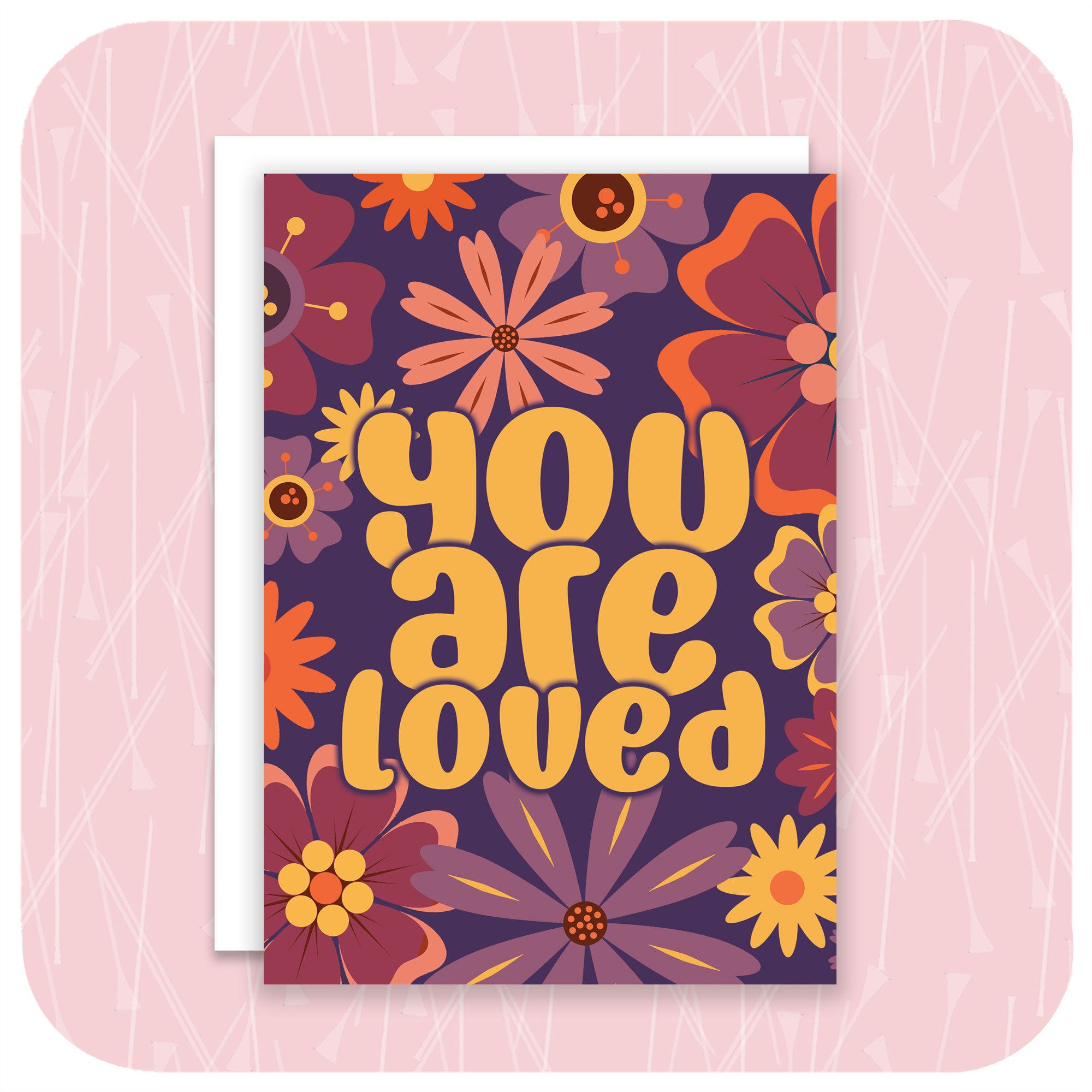 An A6 greetings card featuring 70s / 60s style flowers in pinks, purples & oranges and the words "You Are Loved" with a white envelope sits on a pale pink textured background | The Inkabilly Emporium