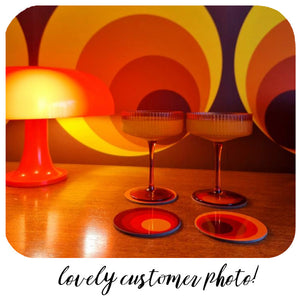 Two cocktail glasses and coasters on a wooden surface with a colourful abstract wall in the background.