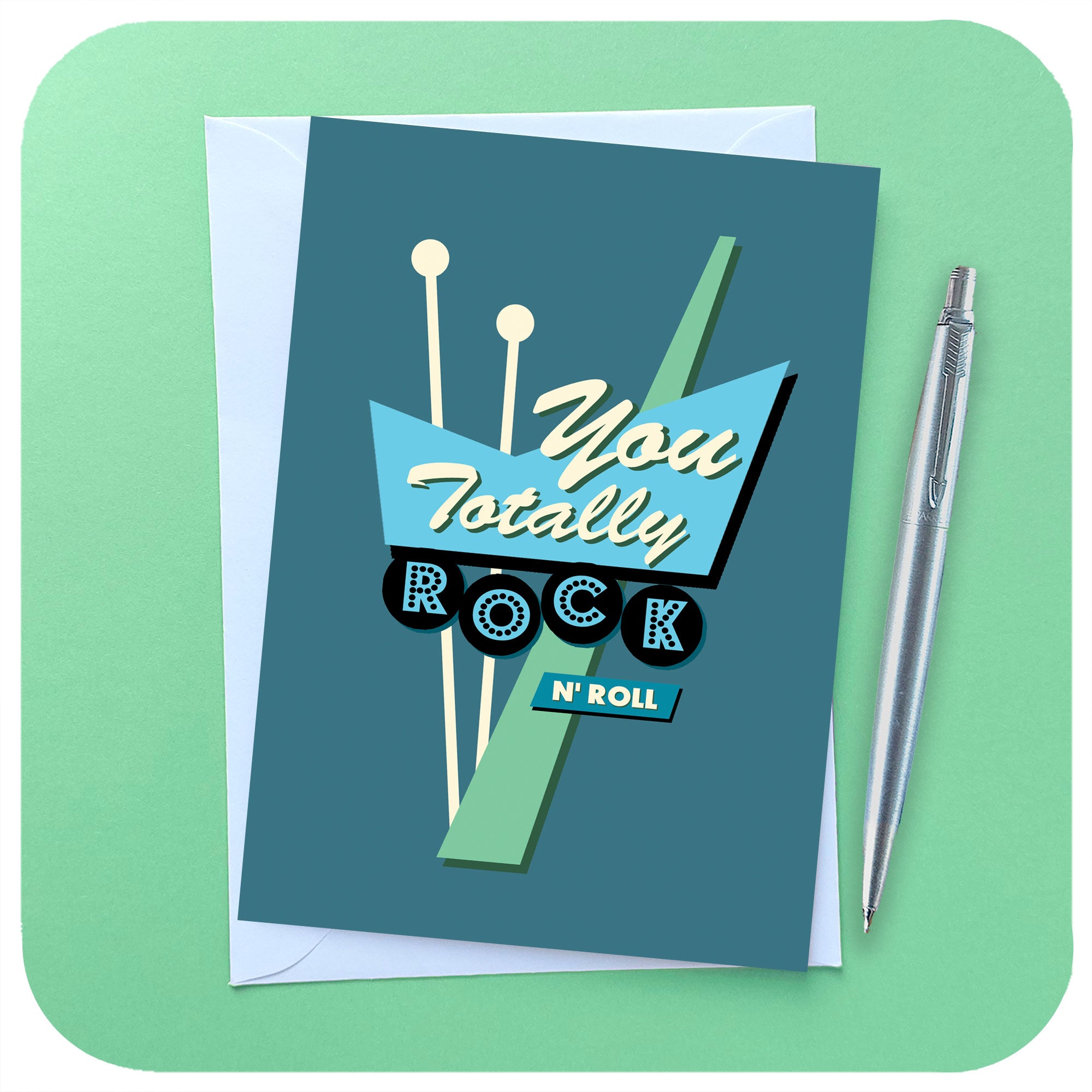 An A6 greetings card featuring a graphic rendering of a 50s style American roadside neon sign in blues and greens, with the words "You Totally Rock n Roll" sits on a white envelope beside a chrome pen on a light green background | The Inkabilly Emporium