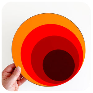 A round 70s style placemat is held against a white background, the placemat has concentric circles in shades of orange and brown | The Inkabilly Emporium