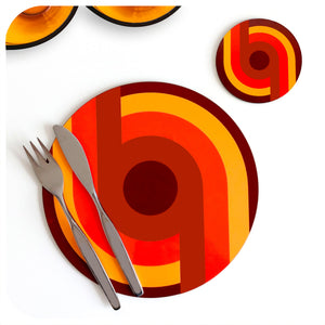 A 70s style round placemat and coaster are set on a white table with mid century cutlery lying across the placemat. The design on the placemat is a supergraphic in oranges and browns | The Inkabilly Emporium