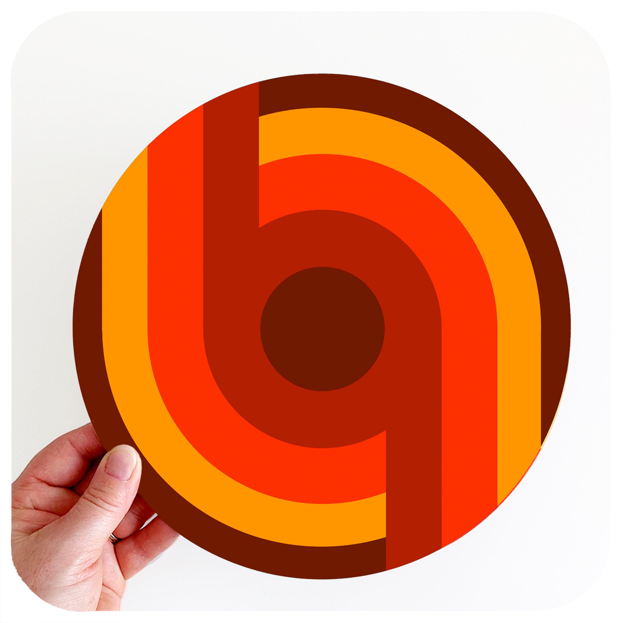 A hand holds up a 70s style round placemat against a white background. The design on the placemat is a supergraphic in oranges and browns | The Inkabilly Emporium