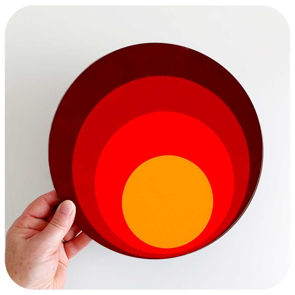 A round 70s style placemat is held against a white background, the placemat has concentric circles in shades of orange and brown | The Inkabilly Emporium
