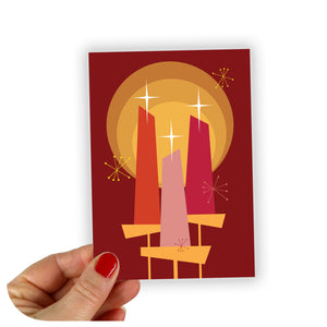 A Christmas Card featuring 3 candles in a mid century modern graphic style is being held by the corner against a white background | The Inkabilly Emporium
