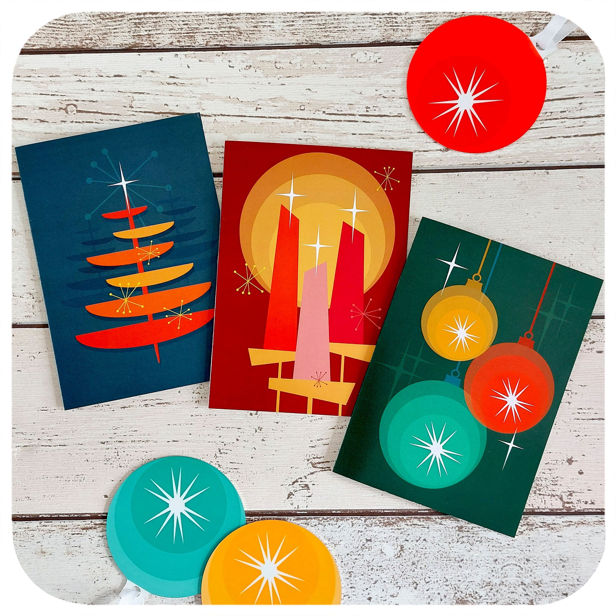 Mid Century Christmas Cards X3, on a whote table with 50s style christmas decorations. Three designs featuring a retro Christmas Tree, Retro baubles and Christmas Candles | The Inkabilly Emporium