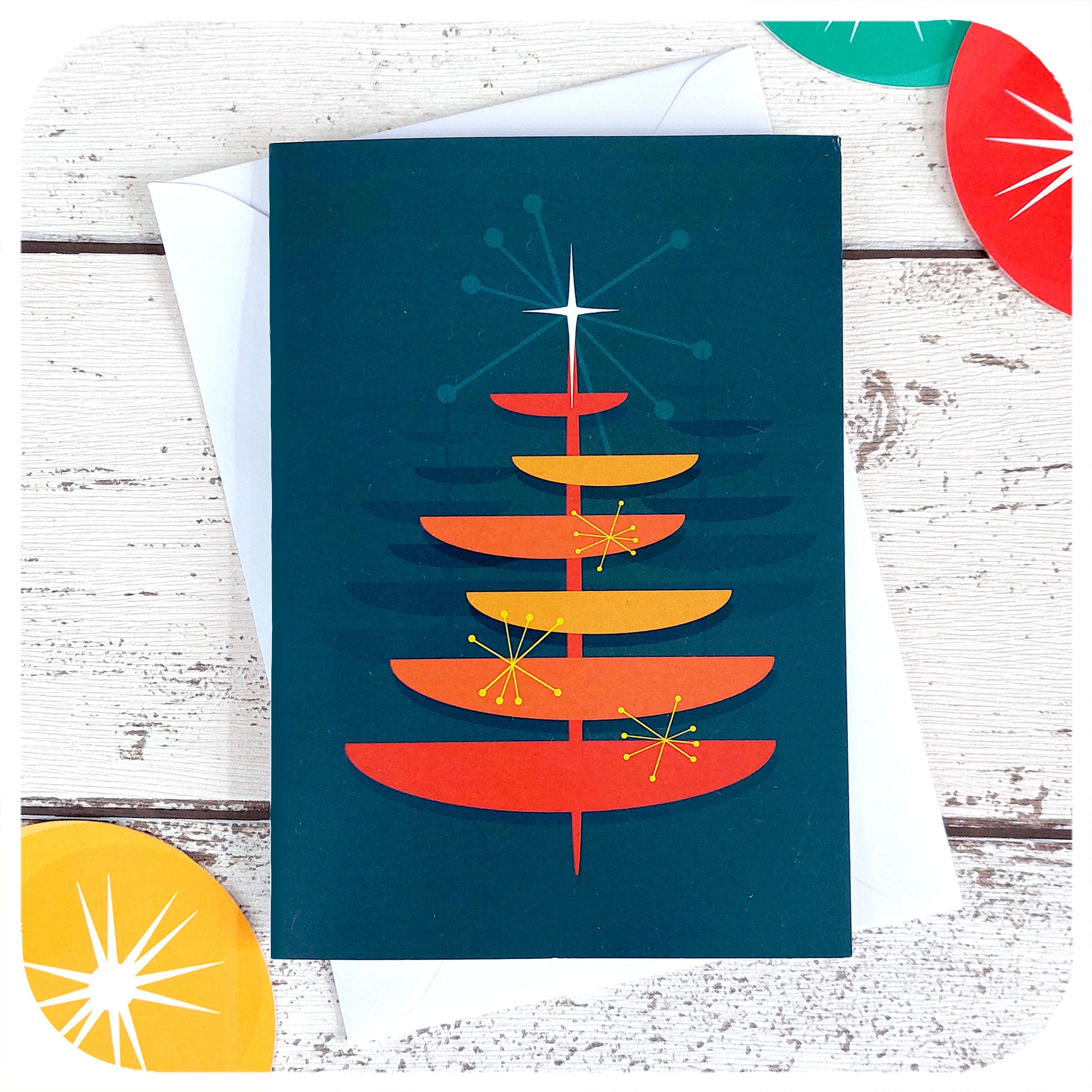 Mid Century style Christmas Card featuring an MCM style orange Christmas Tree on a blue background, with a white envelope, on a white table with 50s style xmas decorations | The Inkabilly Emporium